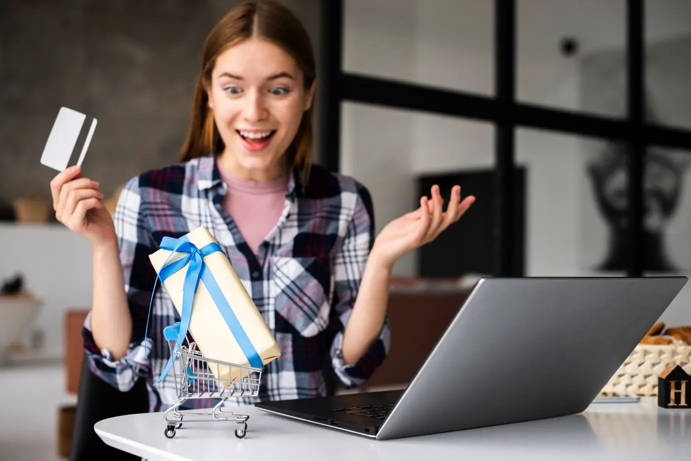 A woman in a checkered shirt holds a credit card and gestures beside a laptop and a small shopping cart with a wrapped gift.