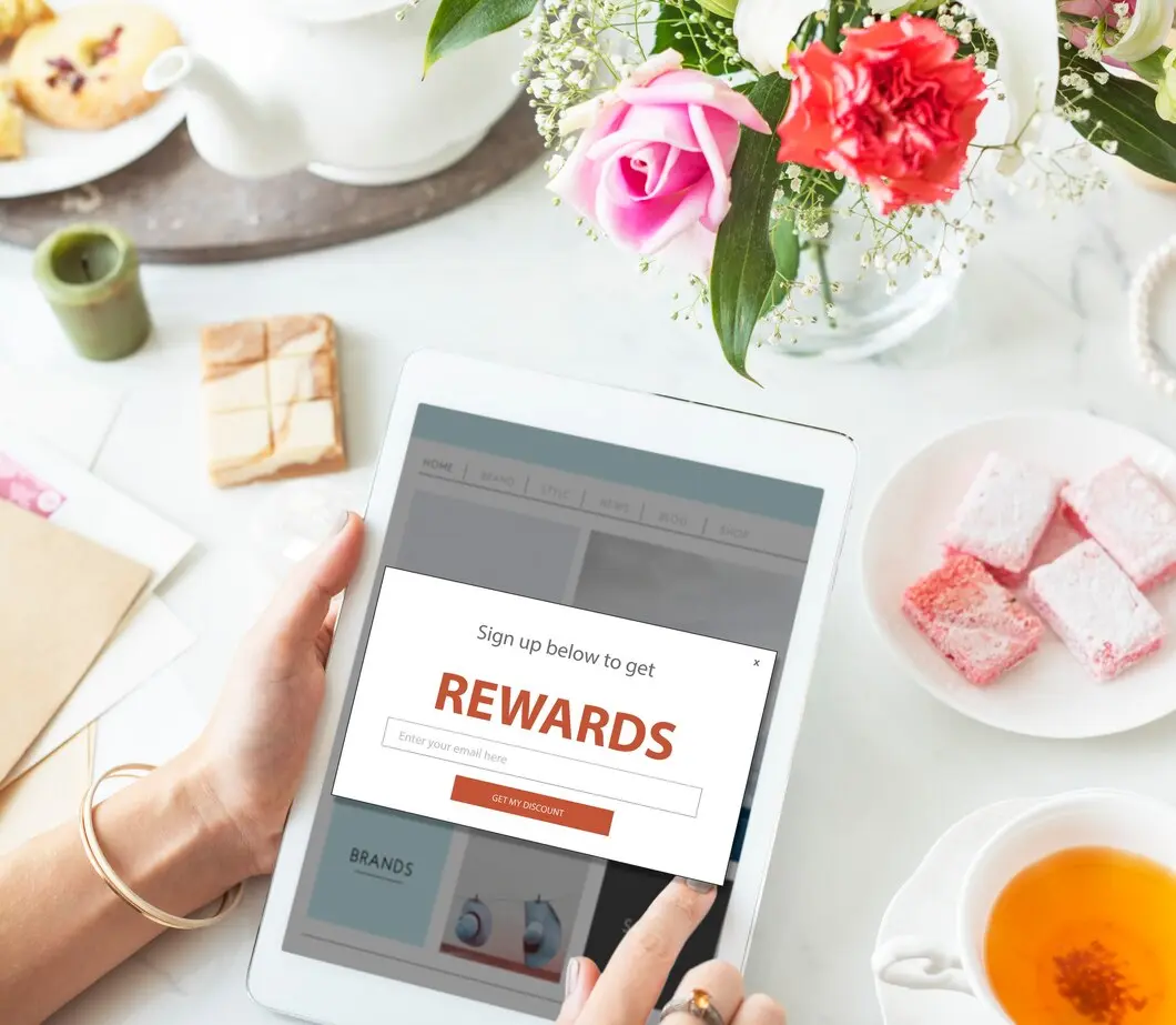 A person holds a tablet displaying a rewards signup form, surrounded by tea, pastries, and fresh flowers on a white table..