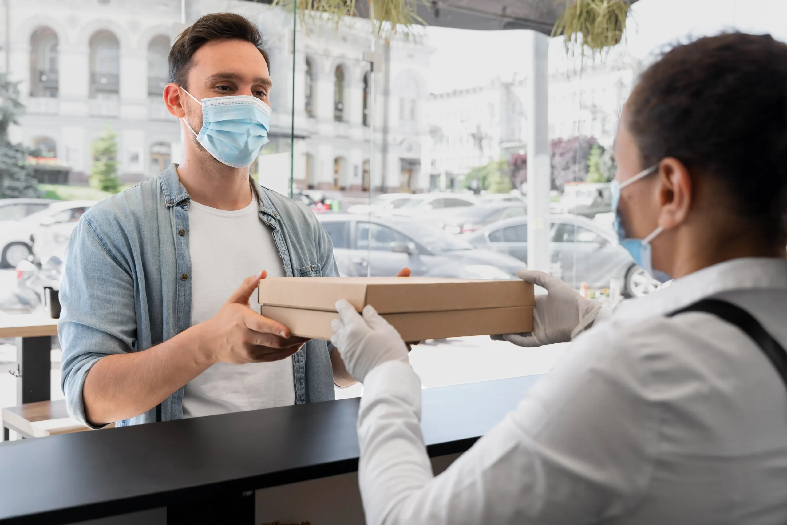A customer receives a package from a masked worker at a modern service counter, with cars parked outside in the background.