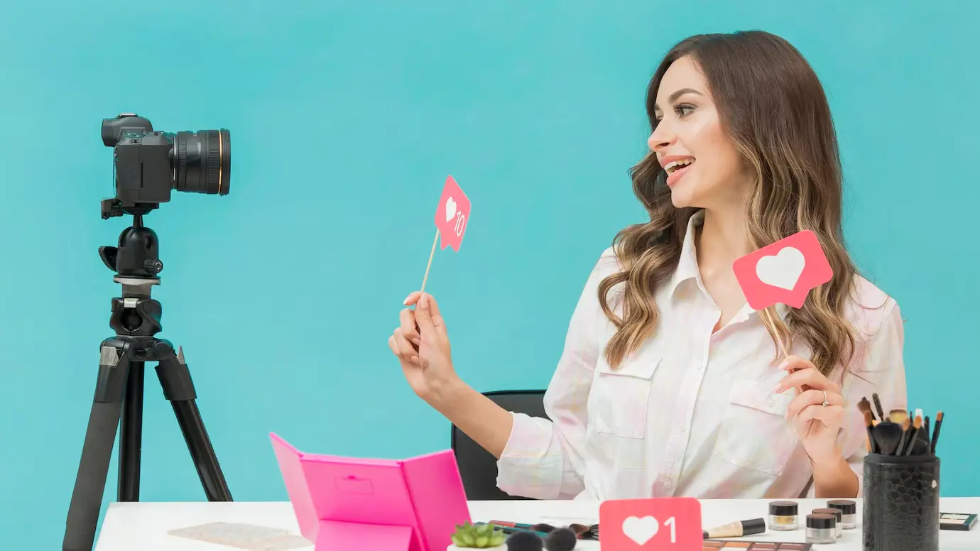 A woman holds pink love sign cards while seated at a table with makeup supplies, smiling at a camera on a tripod against a teal background.